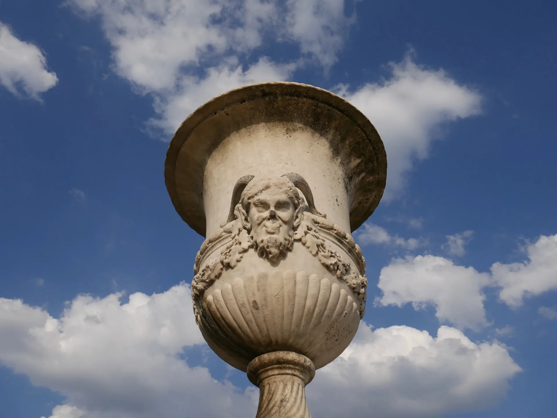 Vase en pierre sculpté avec mascaron à visage barbu sur fond de ciel bleu nuageux